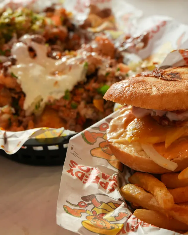 A burger and a plate of loaded nachos side by side, both in baskets lined with Speedy Tacos paper