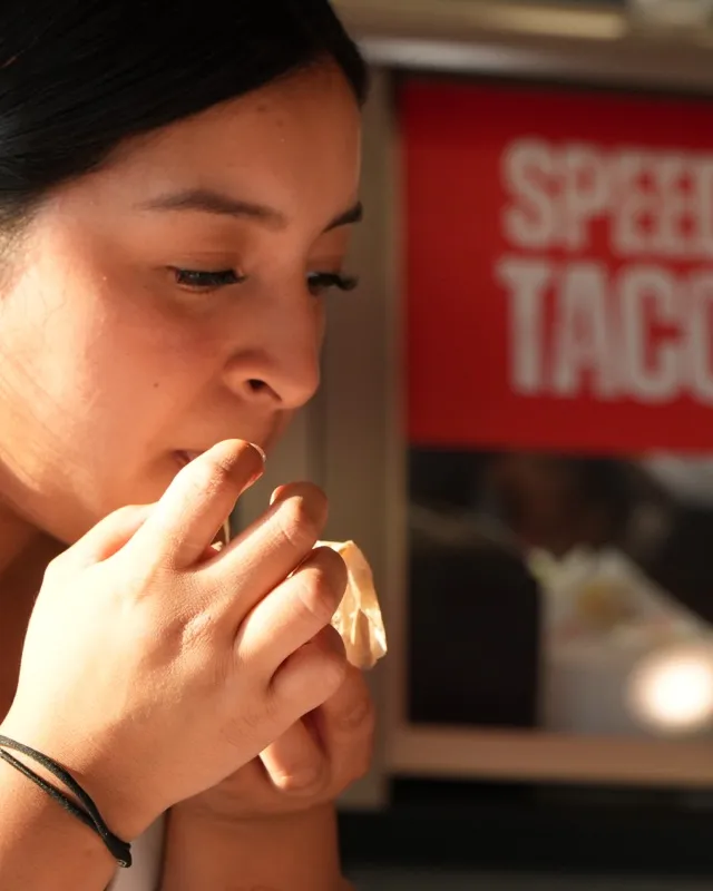 A woman eating a taco with the red Speedy Tacos sign on the wall behind her
