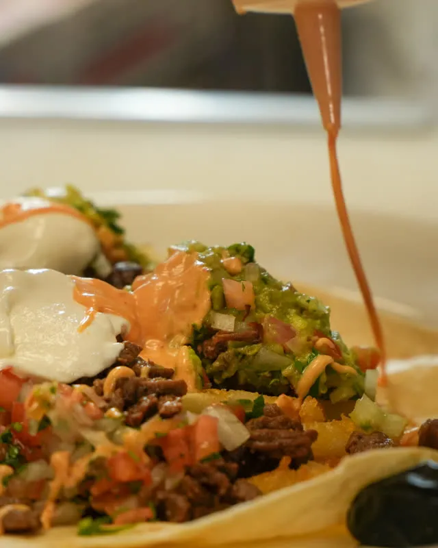 Sauce being poured over a plate of nachos loaded with meat, pico de gallo, and sour cream