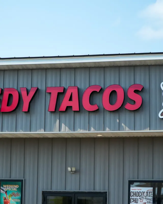The Speedy Tacos sign in red letters on the grey building, the running boy beside it against a blue sky